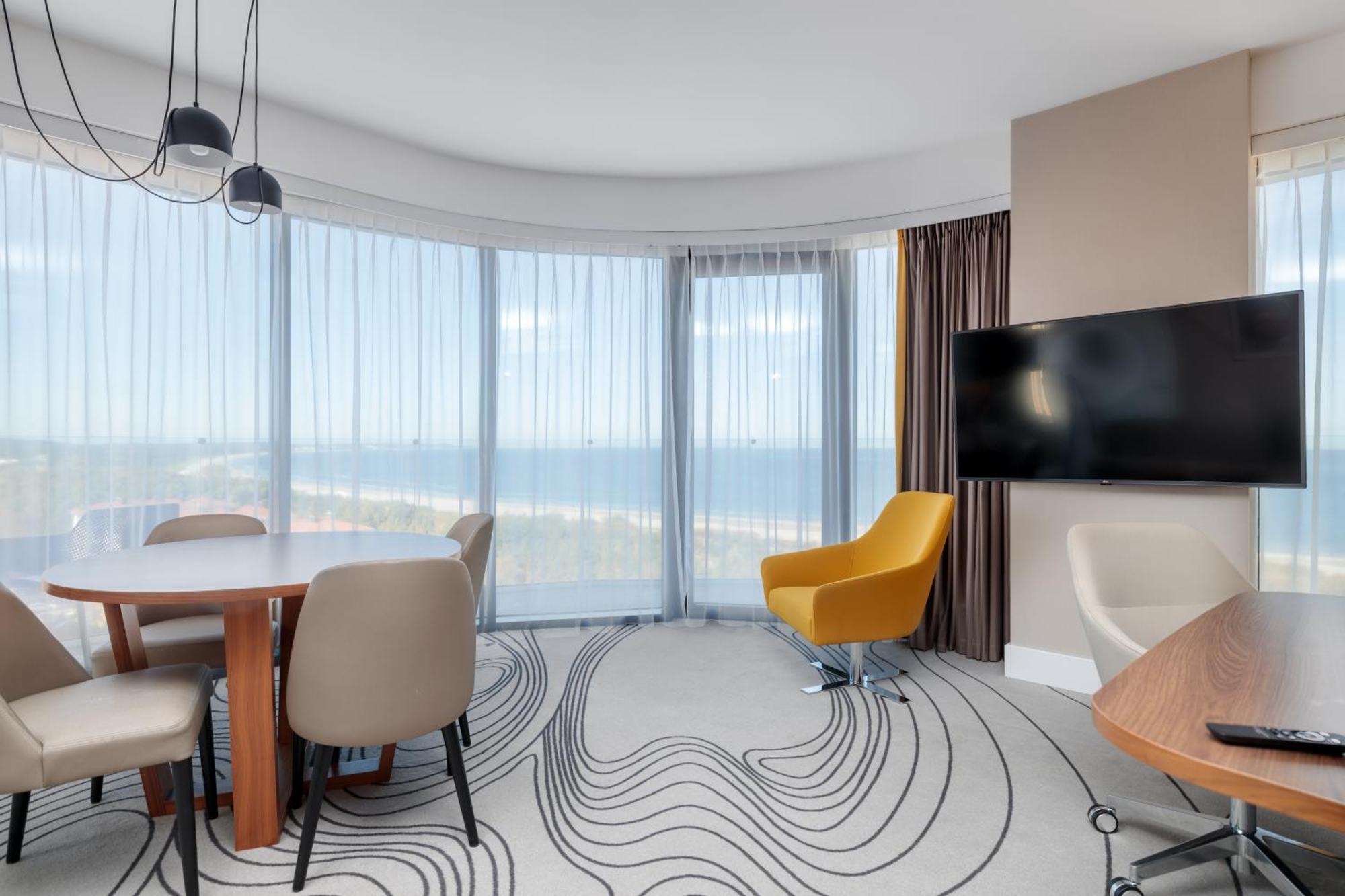 Radisson Blu Resort シフィノウイシチェ エクステリア 写真 The image shows a modern hotel room or suite with a bright, airy atmosphere. There are large windows that allow natural light to fill the space, providing a view of the ocean. The decor includes a round dining table with several chairs, and a stylish