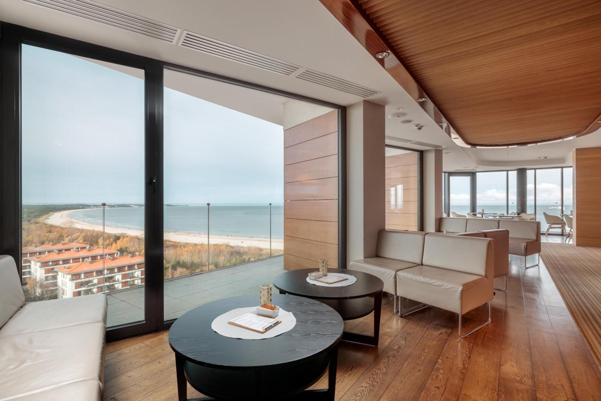 Radisson Blu Resort シフィノウイシチェ エクステリア 写真 The photo shows a modern, elegantly designed interior space featuring large windows that offer a panoramic view of the ocean and coastline. The flooring is wooden, providing a warm atmosphere. There are two round black tables in the foreground with m