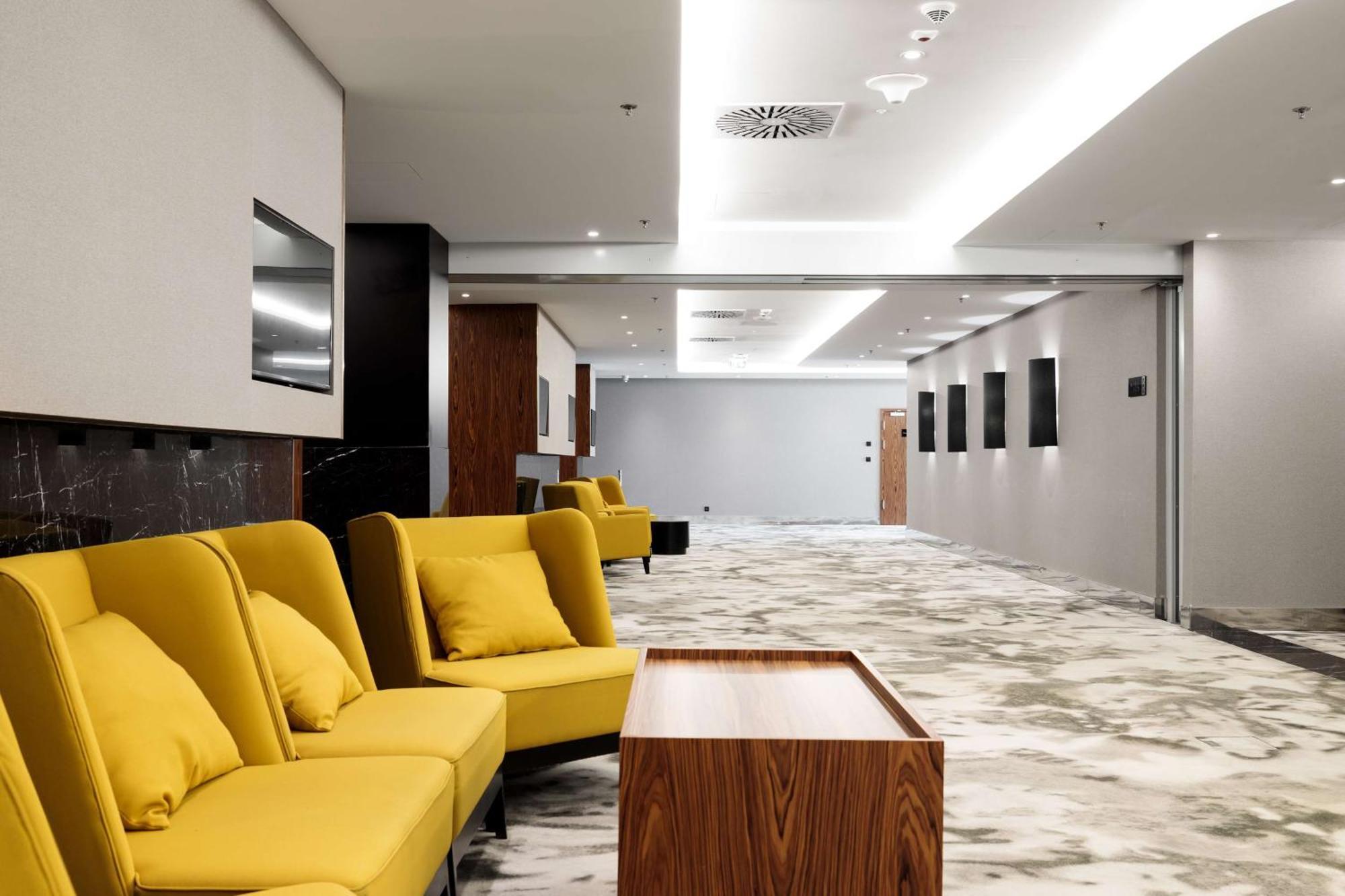 Radisson Blu Resort シフィノウイシチェ エクステリア 写真 The photo shows a modern interior space, likely a lobby or waiting area. There are several yellow sofas with cushions arranged in a casual seating area. In front of the sofas, there is a wooden coffee table. The flooring features a gray and white mar
