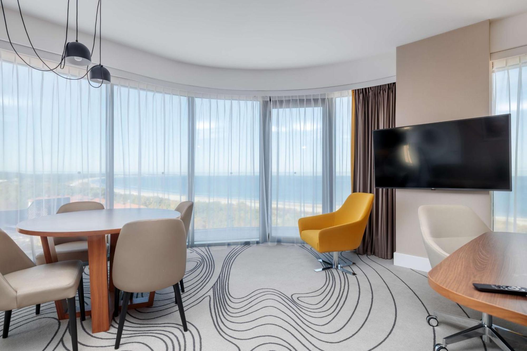 Radisson Blu Resort シフィノウイシチェ エクステリア 写真 The photo shows a modern, stylish interior of a room with large windows that provide a panoramic view. The space features a round dining table surrounded by chairs, including a distinct yellow armchair. There is a flat-screen television mounted on th