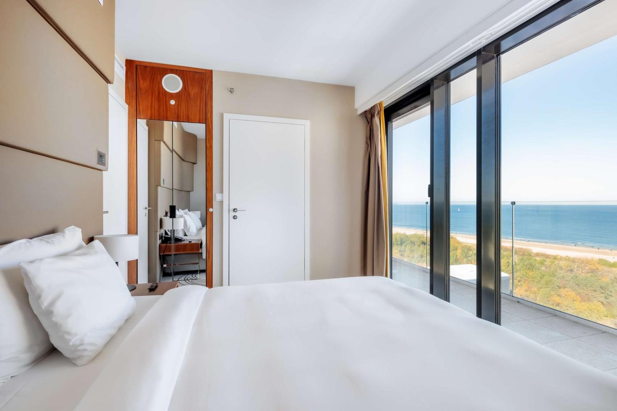 Radisson Blu Resort シフィノウイシチェ エクステリア 写真 The photo shows a modern hotel room with a bright and airy atmosphere. There is a large bed with white bedding positioned near a glass wall that offers a view of the ocean. The room features neutral-colored walls and wooden accents, enhancing its con