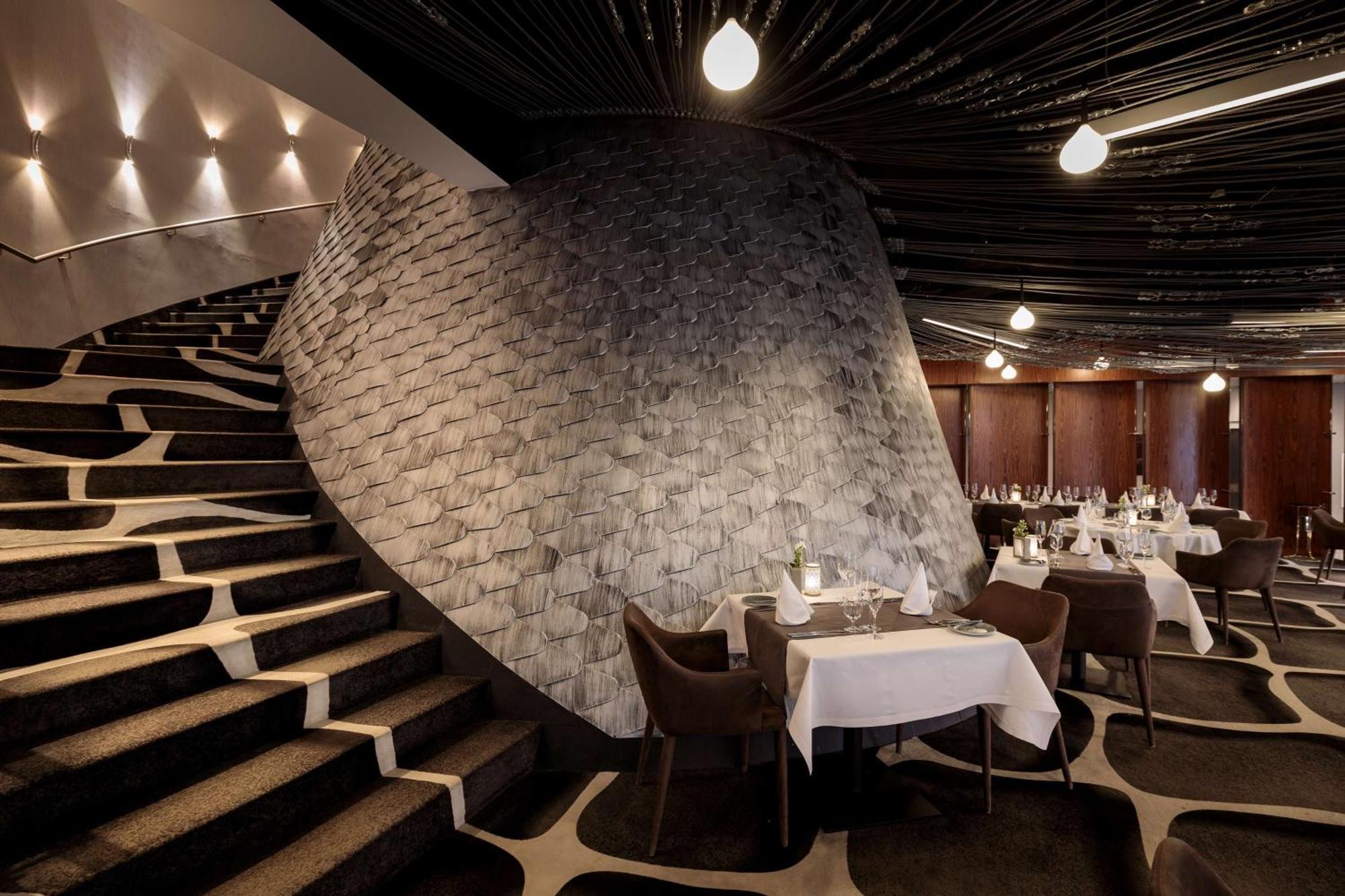 Radisson Blu Resort シフィノウイシチェ エクステリア 写真 The photo shows the interior of a stylish restaurant. In the foreground, there are elegantly set tables with white tablecloths and dark chairs. A unique, curving stone wall adds a distinct architectural feature, and a grand staircase with a textured 