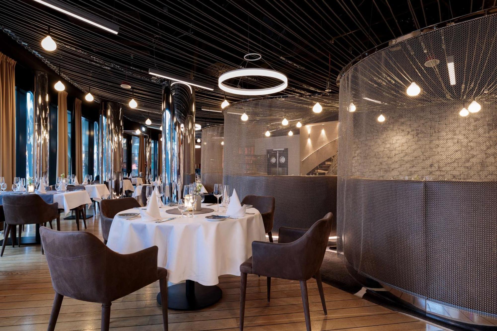Radisson Blu Resort シフィノウイシチェ エクステリア 写真 The image shows a modern restaurant interior that features a stylish design. There are several round tables with white tablecloths. The seating consists of brown, upholstered chairs. The space is illuminated by various hanging lights, including a dis